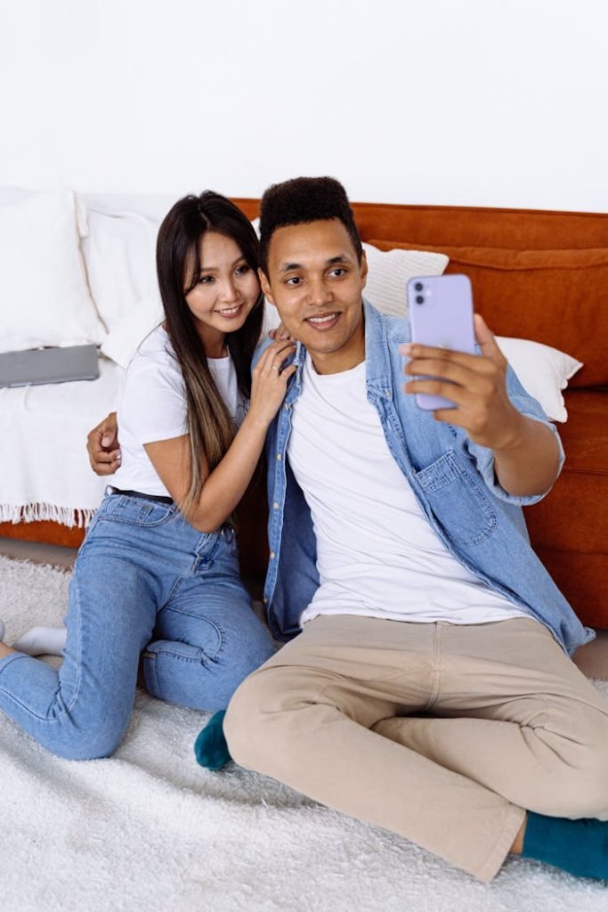 Interracial couple sitting indoors capturing a selfie on a smartphone.